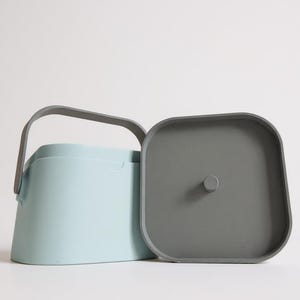 Puede incluir: Un recipiente azul claro con un asa gris y una tapa gris separada. El recipiente tiene una forma rectangular redondeada y un asa que se arquea sobre la parte superior. La tapa es cuadrada con esquinas redondeadas y un peque&ntilde;o pomo.