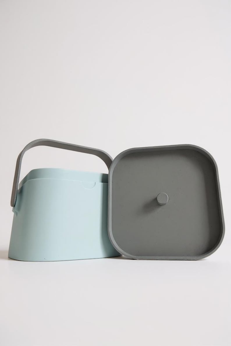 Puede incluir: Un recipiente azul claro con un asa gris y una tapa gris separada. El recipiente tiene una forma rectangular redondeada y un asa que se arquea sobre la parte superior. La tapa es cuadrada con esquinas redondeadas y un peque&ntilde;o pomo.