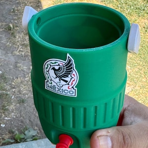 May include: A green cylindrical beverage cooler with a white sticker featuring a black eagle graphic and the text MEXICO. The cooler has two white side tabs, a red bottom base, and a small red circular plug near the bottom of the green body.