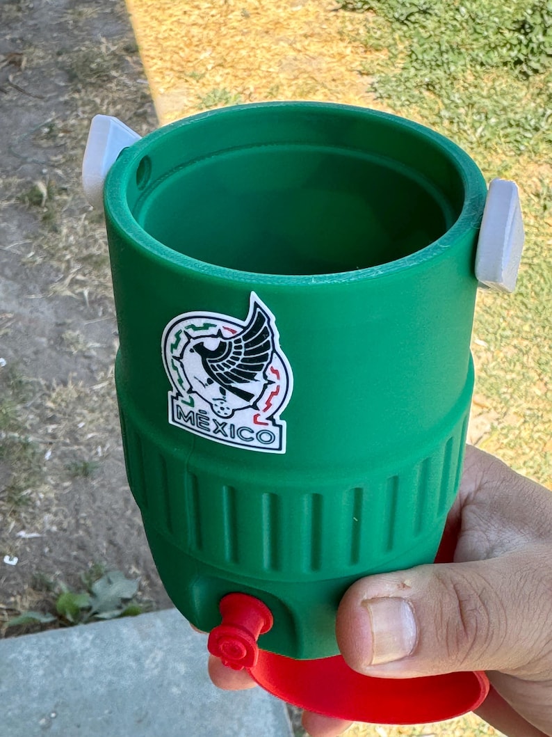 May include: A green cylindrical beverage cooler with a white sticker featuring a black eagle graphic and the text MEXICO. The cooler has two white side tabs, a red bottom base, and a small red circular plug near the bottom of the green body.