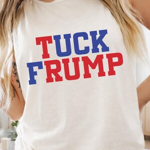 May include: Cream-colored tank top with the text "TUCK FRUMP" in red and blue block letters. The sleeveless shirt is made of a soft material.