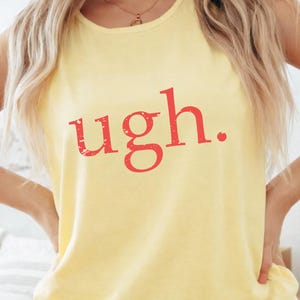 May include: Yellow sleeveless shirt with the word "ugh." printed in red. The shirt is made of a soft material. The model is wearing the shirt with their hands on their hips.