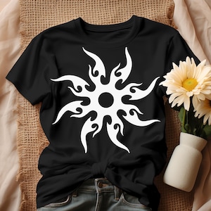 May include: Black t-shirt featuring a white tribal sunburst design. The graphic has a central circle with flame-like shapes radiating outwards. The shirt is paired with denim jeans and a small vase of flowers.