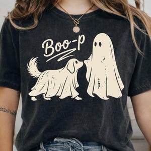 May include: A charcoal grey short-sleeve t-shirt featuring a cream-colored graphic of a dog wearing a ghost costume standing next to a ghost. The text "Boo-p" is printed above the dog in a stylized font. The shirt has a faded, vintage-style wash.