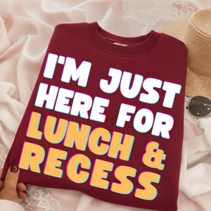 Può includere: Una felpa girocollo color bordeaux con il testo bianco e giallo "I'M JUST HERE FOR LUNCH & RECESS" in un carattere audace e stratificato. L'indumento presenta un colletto a coste e maniche lunghe, presentato come un capo disteso.