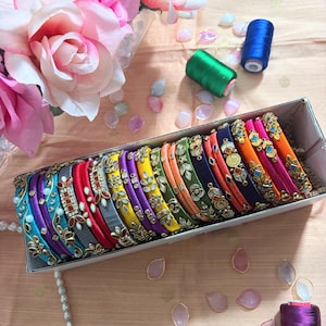 May include: A rectangular box filled with a collection of multi-colored silk thread bangles, each featuring intricate gold-tone metal embellishments and small inset stones. The bangles are arranged in a row, showcasing a vibrant spectrum of colors.