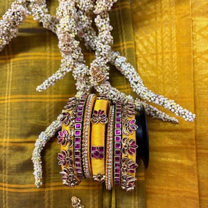 May include: A set of yellow silk-wrapped bangles featuring metallic accents with pink stone inlays and floral motifs. The bangles are arranged in a stack, showcasing intricate gold-toned metalwork and small clear crystals, set against a mustard yellow fabric.
