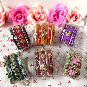 May include: Six sets of bangles wrapped in colorful silk thread, each featuring intricate metallic embellishments, small faux pearls, and decorative stones. The bangles are arranged in groups and encased in clear plastic protective packaging.