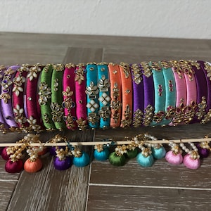 May include: A collection of multi-colored silk thread bangles featuring intricate gold-tone metal embellishments with clear stones, arranged in a row, accompanied by a set of matching small hoop earrings with dangling silk thread tassels.
