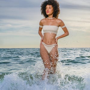 May include: A cream-colored crochet bikini set, including a bandeau top and high-waisted bottoms. The model is standing in the ocean, with waves splashing around them. The set is accessorized with a shell necklace and arm cuffs. The sky is a mix of blue and yellow.