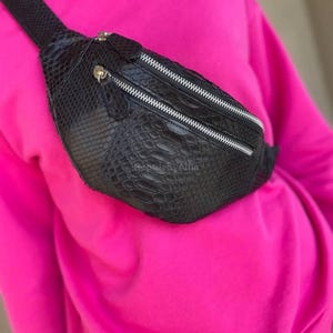 Puede incluir: Riñonera negra con un patrón texturizado similar a la piel de serpiente. La bolsa tiene dos compartimentos con cremalleras plateadas. La correa también es negra y parece ajustable. La riñonera se muestra sobre un fondo rosa brillante.