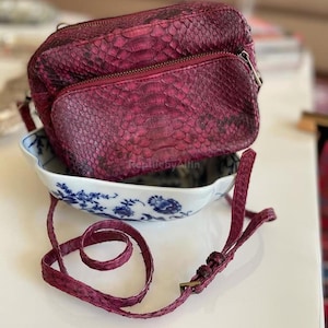 Könnte beinhalten: Burgunderfarbene Umhängetasche mit Schlangenmuster. Die Tasche hat eine Reißverschluss-Vordertasche und einen langen, verstellbaren Riemen. Sie liegt in einer weißen Schale mit blauem Blumenmuster.