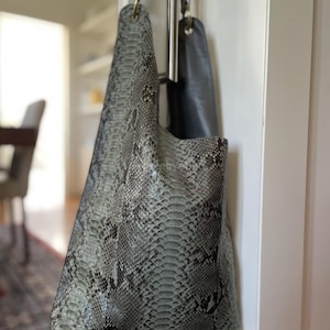 Może przedstawiać: Szara i czarna torba typu tote z wzorem skóry węża z paskiem na ramię. Torba wisi na metalowym drążku przymocowanym do białych drzwi. Wnętrze torby jest w jednolitym szarym kolorze.