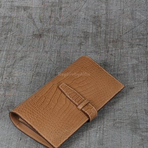 May include: A tan leather wallet with a crocodile skin texture. The wallet is rectangular with a flap and a strap closure. The wallet is closed and lying on a grey surface.