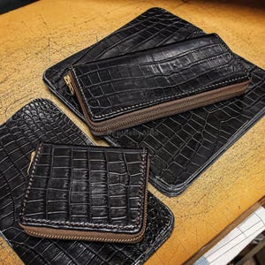 May include: A selection of black leather wallets and cardholders with a crocodile skin effect. The wallets feature brown zippers and stitching. The items are displayed on a wooden surface.
