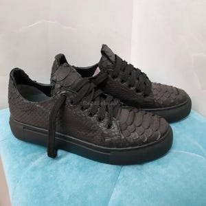 Può includere: Un paio di sneakers nere con un motivo strutturato e squamoso. Le scarpe hanno lacci e suole neri. Le sneakers sono esposte su una superficie blu.