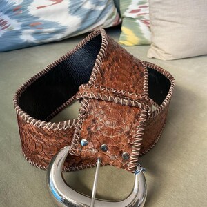 Peut inclure: Large ceinture marron avec un motif peau de serpent et une grande boucle argentée. La ceinture a des coutures blanches sur les bords et un intérieur noir. La boucle est de couleur argent poli.