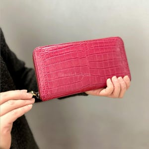 May include: A bright pink wallet with a crocodile skin effect. The rectangular wallet has a zip closure and a gold-coloured zip pull. The wallet is held open, displaying its design and colour.