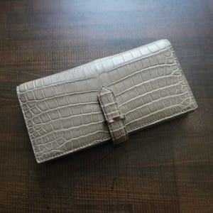May include: A light grey wallet with a crocodile skin texture. It features a strap closure with a silver-coloured metal ring. The wallet is rectangular and rests on a dark brown wooden surface.