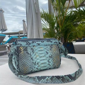 May include: A turquoise and black snakeskin-patterned crossbody bag with a silver-tone chain and a long strap. The bag has a rectangular shape with a zipper closure and a matching strap. The bag is sitting on a white surface.