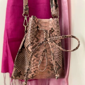 Puede incluir: Bolso tipo cubo con estampado de piel de serpiente en tonos marrón y beige. El bolso tiene un cierre de cordón y una correa larga. El fondo presenta una bufanda rosa brillante. El bolso mide unos 20 cm de alto.