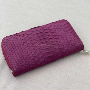 May include: A plum-coloured, zipped wallet with a textured, scaly pattern. The wallet is rectangular and features a silver-toned zip pull. The material appears to be reptile skin, possibly snakeskin. The overall design is sleek and modern.