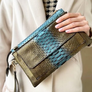 May include: A clutch with a snakeskin pattern in shades of teal and gold. The clutch has a flap closure and a wrist strap. The texture of the snakeskin is visible, and the colors create a striking contrast. The bag is held by a person.