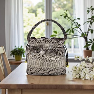 May include: A snakeskin-patterned handbag with a drawstring closure and a top handle. The bag is primarily gray and white with black accents. Gold-colored hardware is visible on the drawstring ends and handle attachments. The bag is sitting on a wooden table.
