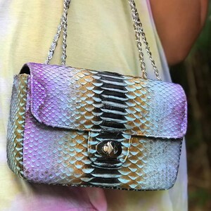 May include: A small, rectangular handbag with a flap closure and silver chain strap. The bag features a snakeskin pattern in shades of purple, gold, black, and blue. The clasp is silver-toned.