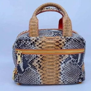 Op de afbeelding: Een vierkante handtas met een slangenprint in bruin, grijs en zwart. De tas heeft een gouden rits, een handvat aan de bovenkant en gouden hardware. De binnenkant is rood.