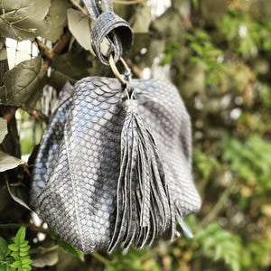 May include: A gray, textured handbag with a tassel and a strap. The bag has a scale-like pattern and a gold-colored ring connecting the strap to the bag. The background is blurred with green foliage.