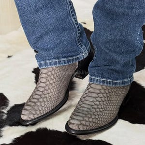 Può includere: Un paio di stivali da cowboy grigi con un motivo strutturato simile alla pelle di serpente. Gli stivali hanno suola e tacco neri e sono indossati con jeans blu. Gli stivali sono di colore neutro.