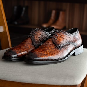 Può includere: Un paio di scarpe eleganti in pelle marrone con un motivo strutturato a scaglie. Le scarpe hanno lacci e un tacco basso, appoggiate su una sedia chiara. Lo sfondo mostra altre scarpe su una mensola.