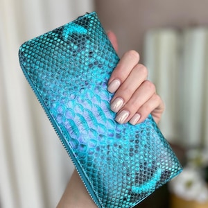 May include: A teal and blue snakeskin-textured wallet with a zipper closure. The wallet has a rectangular shape and is held by a hand. The color transitions from teal to blue and purple. The wallet appears to be made of leather.