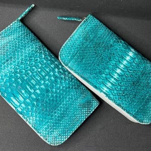 May include: Two turquoise-colored, rectangular zippered pouches with a snakeskin pattern. The pouches are made of a textured material and have a white trim. The zipper pulls are the same turquoise color as the pouches.