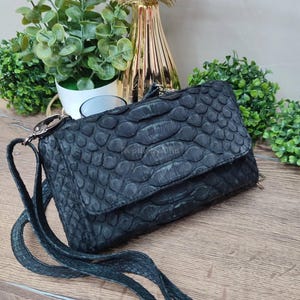 May include: A black, textured leather crossbody bag with a flap closure. The bag features a scale-like pattern and a long, thin strap. The hardware is silver-toned. The bag is displayed on a wooden surface.