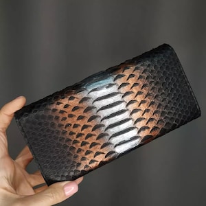 May include: A black and brown snakeskin wallet. The wallet has a rectangular shape and is held by a hand. The snakeskin pattern is visible on the front of the wallet. The wallet has a dark brown and black colour scheme.