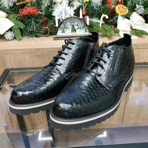 Può includere: Paio di stivali in pelle nera con motivo a pelle di serpente. Gli stivali hanno un design stringato, una cerniera laterale e una suola bianca e nera. Gli stivali sono esposti su una superficie di vetro.