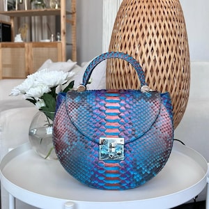 Puede incluir: Un bolso redondo con un vibrante estampado de piel de serpiente en tonos azules, morados y rojos. El bolso tiene un asa superior y un cierre de broche plateado. El bolso está sobre una mesa blanca.