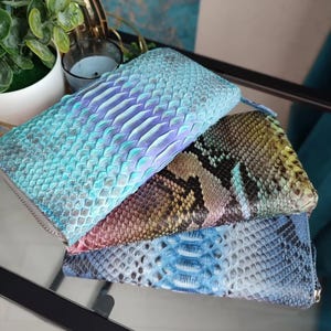 May include: Three snakeskin wallets in shades of blue, purple, brown, and yellow. The wallets have a textured finish and a zip closure. The wallets are stacked on top of each other, and appear to be made of genuine leather. The wallets are on a reflective surface.