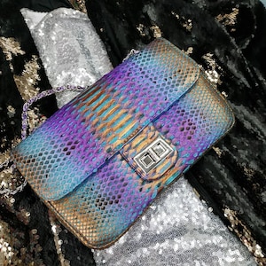 Può includere: Borsa rettangolare con chiusura a patta, caratterizzata da un vivace motivo a pelle di serpente iridescente nei toni del viola, blu e oro. La borsa ha una tracolla a catena argentata e una chiusura argentata. È un accessorio di moda.