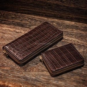 May include: Two brown leather wallets with a crocodile skin texture are displayed on a wooden surface. The larger wallet is rectangular and zippered, while the smaller one is square and also zippered. Both have gold-coloured zippers.