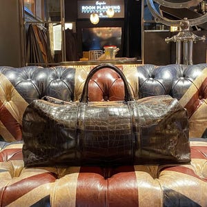 Puede incluir: Una bolsa de lona de cuero marrón con textura de piel de cocodrilo descansa sobre un sofá de cuero con estampado de la bandera británica. La bolsa tiene un asa superior y un cierre de cremallera. El fondo incluye un espejo y un letrero que dice "ROOM PLANNING".