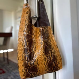 May include: A large, open-top handbag with a snakeskin pattern in shades of brown and tan. The bag has a dark brown leather interior and a single strap. The bag is hanging from a metal hook.