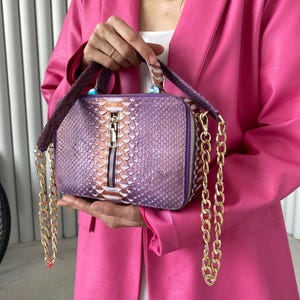 May include: A rectangular, purple handbag with a snakeskin pattern and a gold zipper. The bag has a gold chain strap and a handle. The bag is held by a person wearing a pink blazer.