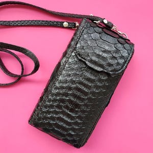 May include: Black leather crossbody bag with a textured, scaly pattern. The bag has a flap closure and a long, adjustable strap. The bag is shown against a bright pink background. The bag is approximately 7 inches long.