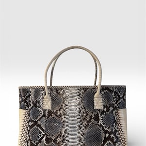 Könnte beinhalten: Rechteckige Handtasche mit Schlangenmuster in Grau, Schwarz und Creme. Die Tasche hat zwei cremefarbene Griffe und cremefarbene Akzente an den Seiten. Die Tasche ist im Shopper-Stil gehalten.