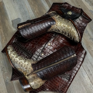 Può includere: Un gilet in pelle marrone e stivali da cowboy abbinati. Il gilet presenta un motivo strutturato a forma di coccodrillo con bottoni color oro. Gli stivali presentano un design a pelle di serpente sulla tomaia e una chiusura con cerniera.