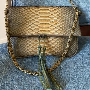Könnte beinhalten: Eine Handtasche mit Schlangenmuster und einem goldfarbenen Kettenriemen. Die Tasche hat eine Klappe mit Reißverschluss und dekorativen grünen Quasten. Das Schlangenmuster ist in Braun- und Beigetönen gehalten.