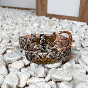 Könnte beinhalten: Ein aufgerollter Schlangenleder-Gürtel mit einer goldfarbenen Schnalle. Der Gürtel weist ein Muster aus braunen, beigen und schwarzen Schuppen auf. Die Schnalle ist ein einfaches, rechteckiges Design. Der Gürtel liegt auf einem Bett aus weißen Steinen.